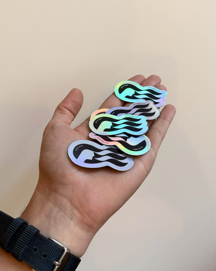 Hand holding several holographic, wave-shaped stickers with a black mermaid silhouette and the text 'SEA PRINCESS' around the edge, rainbow sheen across the surface
