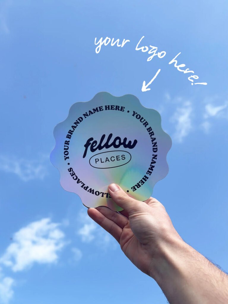 Hand holding a rainbow scalloped sticker that reads 'fellow places' with placeholder 'your brand name' text around the edge, against a blue sky (logo mockup).