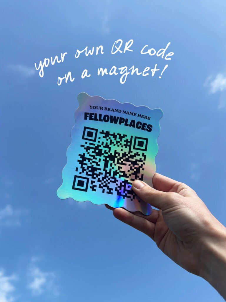 Hand holding a rainbow gradient QR code magnet against a bright blue sky, with the caption “your own QR code on a magnet!”.